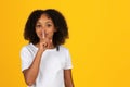 Smiling adolescent african american girl in white t-shirt, puts finger to lips, makes shhh sign Royalty Free Stock Photo
