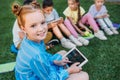 smiling adorable schoolgirl using tablet while sitting on grass