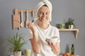 Smiling adorable young woman posing in kitchen with towel on head applying facial anti aging mask enjoying skin care procedures