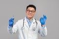 Smiling adult asian doctor in white coat, glasses, protective gloves hold syringe, show ok