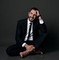 Smiling adult bearded man in black office suit sits barefooted on floor with legs crossed holding hand at head