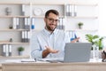 Smiling adult caucasian male in glasses gestures and looks at laptop in living room or office interior Royalty Free Stock Photo