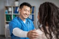 Smiling adult chinese male doctor calms and supports young african american woman patient in clinic