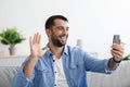 Smiling adult european guy with beard makes online call and waves hand to webcam at home interior