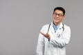 Smiling adult korean man therapist in white coat, glasses with stethoscope shows hand on empty space