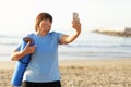 Smiling adult woman in training wear holding yoga mat make photo or have video call with smartphone over sea nature Royalty Free Stock Photo