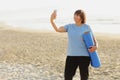 Smiling adult woman in training wear holding yoga mat make photo or have video call with smartphone over sea nature Royalty Free Stock Photo