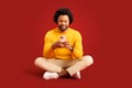 Smiling african american curly man with smartphone in his hands sitting in lotus pose isolated on red Royalty Free Stock Photo