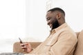 Smiling african american man browsing news or social media feed on phone while sitting on sofa at home. Rest and Royalty Free Stock Photo
