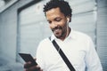 Smiling African American man in headphones standidng in sunny street listening to songs on his cell phone, using Royalty Free Stock Photo