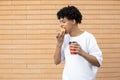 Smiling African-American man holding a cup of coffee and biting an orange donut Royalty Free Stock Photo