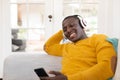 Smiling african american man at home sitting on couch wearing headphones and looking at smartphone Royalty Free Stock Photo