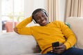 Smiling african american man at home sitting on couch wearing headphones and looking at smartphone Royalty Free Stock Photo