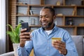 A smiling African American man is at home, sitting on the sofa, holding a credit card and using a mobile phone. Close-up Royalty Free Stock Photo