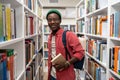 Smiling african american man student holds book in university library between bookshelves. Royalty Free Stock Photo