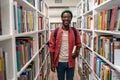 Smiling african american man student holds book in university library between bookshelves. Royalty Free Stock Photo