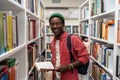 Smiling african american man student holds book in university library between bookshelves. Royalty Free Stock Photo