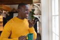 Smiling african american man in talking on smartphone holding coffee looking out of kitchen window Royalty Free Stock Photo