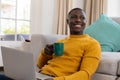 Smiling african american man using laptop sitting on floor by couch at home drinking coffee Royalty Free Stock Photo