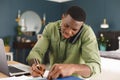 Smiling african american man working at home in bedroom using laptop, talking on phone and writing Royalty Free Stock Photo