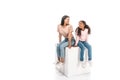 African american mother and daughter looking at each other while sitting on white cube on white background