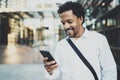 Smiling African American while walking at the city holding mobile phone in hands and checking email.Blurred background. Royalty Free Stock Photo