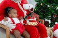 Smiling African child girl baby eating snacks during sitting on Santa Claus chair with decorative Christmas tree blurred Royalty Free Stock Photo