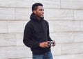 Smiling african man photographer with vintage film camera looking away wearing black hoodie while walking on city street Royalty Free Stock Photo