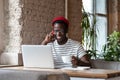 Smiling African man talking on phone, holding cup of coffee, remotely online work on laptop in cafe Royalty Free Stock Photo