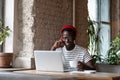 Smiling African man talking on phone, holding cup of coffee, remotely online work on laptop in cafe Royalty Free Stock Photo