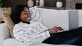 Smiling african woman student girl freelancer user lying on couch using laptop typing message online chat with friends Royalty Free Stock Photo
