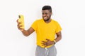 Smiling Afro American student or freelancer standing near the wall in trendy yellow t-shirt, having online call with Royalty Free Stock Photo