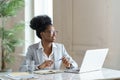 Smiling Afro woman in blazer wear glasses working at laptop computer at home office, looking away.