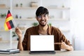 Smiling arab guy student showing flag of Germany and laptop