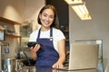 Smiling asian barista, girl with card terminal, payment machine and laptop, standing in cafe, processing payment for Royalty Free Stock Photo