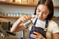 Smiling asian barista girl, making coffee, pouring steamed milk into cappuccino, doing latte art in cup, working in cafe Royalty Free Stock Photo