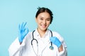 Smiling asian doctor, female physician touching stethoscope, showing okay, ok sign in approval, blue background