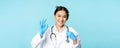 Smiling asian doctor, female physician touching stethoscope, showing okay, ok sign in approval, blue background