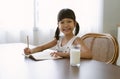 Smiling Asian elementary school girl writing or drawingcoloring pages in notebook and looking at camera while sitting behind desk Royalty Free Stock Photo