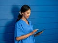 Smiling Asian female doctor or nurse in blue scrubs suit using smart mobile phone while standing on blue background. Royalty Free Stock Photo