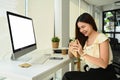 Smiling asian female worker sitting in front of blank computer display at office desk and eating sandwich for lunch Royalty Free Stock Photo