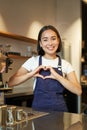 Smiling asian girl barista, shows heart sign, loves making coffee an serving clients, standing in uniform behind counter