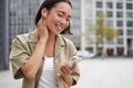 Smiling asian girl listens music in wireless headphones, looks at her phone, choosing music or podcast. Young woman Royalty Free Stock Photo