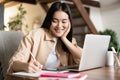 Smiling asian girl student studying from home, online learnig, writing with pen in notebook, working with laptop