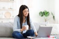 Smiling asian girl unwinding with laptop and coffee on sofa at home