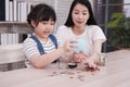 Smiling Asian little asian girl child is putting coins into piggy bank for saving money for the future with mother on wooden table Royalty Free Stock Photo