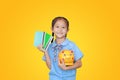 Smiling Asian little girl in school uniform holding account book and piggy bank isolated on yellow background. Schoolgirl with Royalty Free Stock Photo