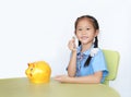 Smiling Asian little girl in school uniform sitting on table show putting coin into piggy bank isolated on white background. Royalty Free Stock Photo