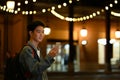 Smiling asian man checking his mobile phone while standing in city avenue with blurred night street lights background Royalty Free Stock Photo