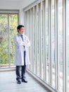 Smiling Asian man doctor portrait in white coat standing alone with crossed arms near glass windows in medical office. Royalty Free Stock Photo